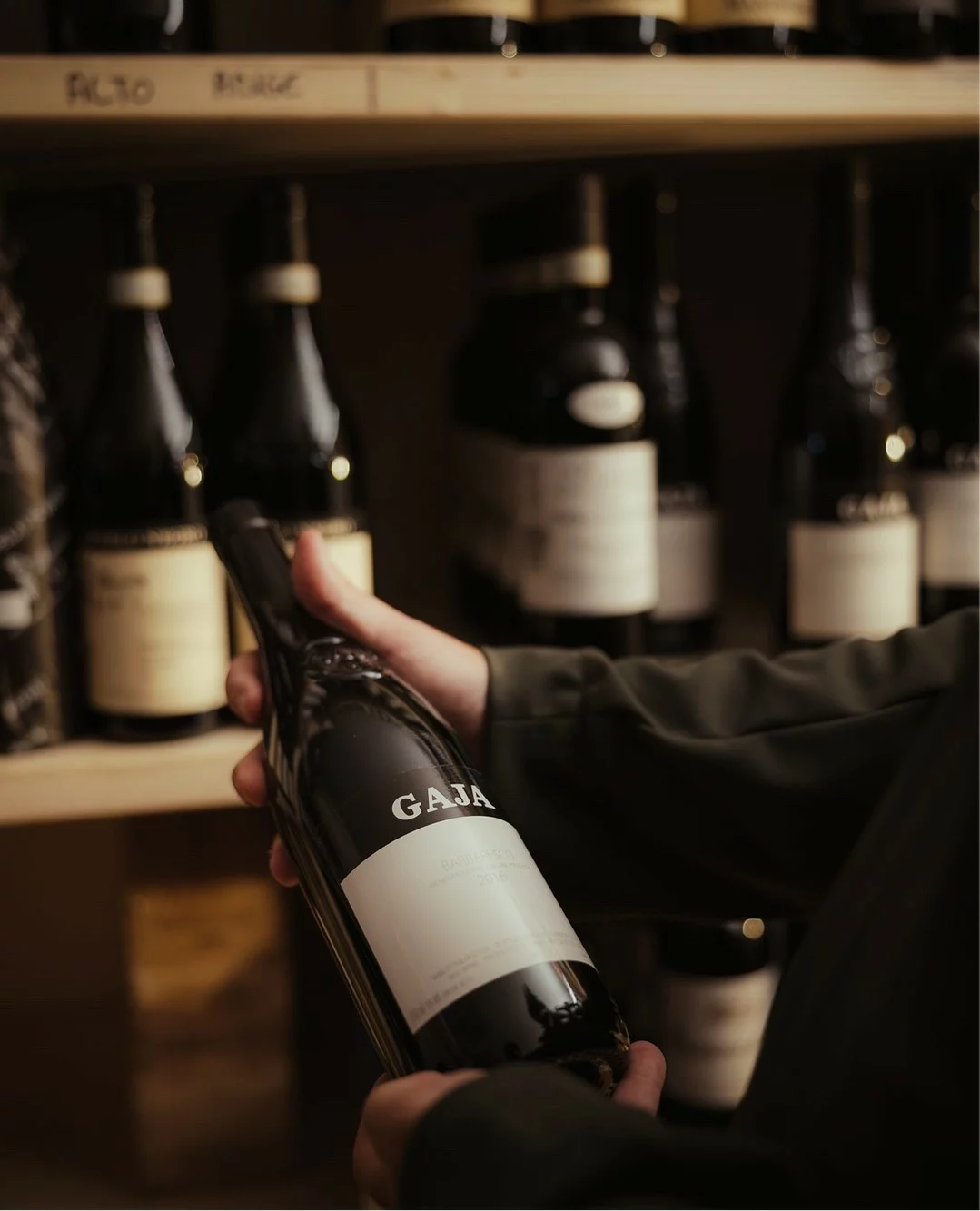 Mani tengono bottiglia di vino Gaja con altre bottiglie sullo sfondo.