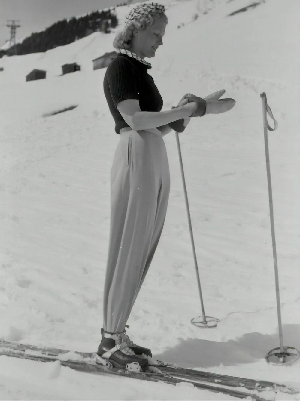 Foto in bianco e nero di donna sugli sci con racchette nella neve infila i guanti.