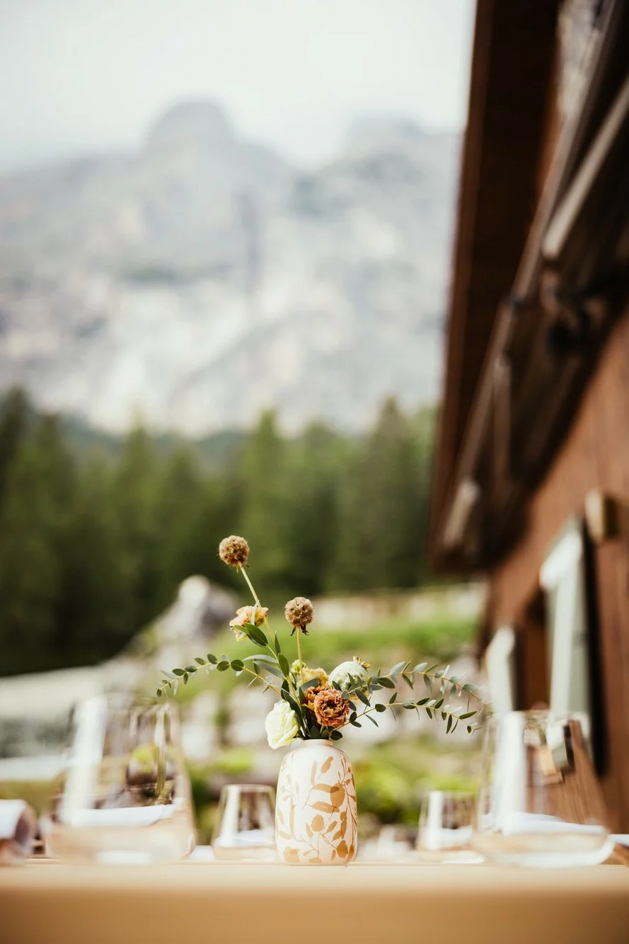 Tavolo esterno apparecchiato con piccolo vaso con fiori di campo con alberi e montagna sullo sfondo.